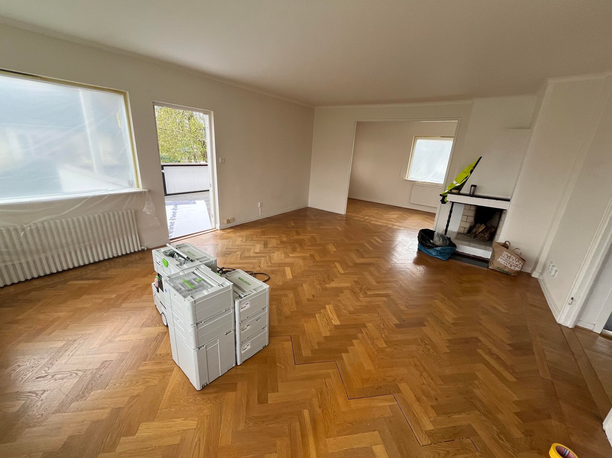 Jasne wnętrze z parkietem w jodełkę, zabezpieczonym oknem folią malarską i narzędziami Festool. Widoczny kominek i przejście do innego pomieszczenia.