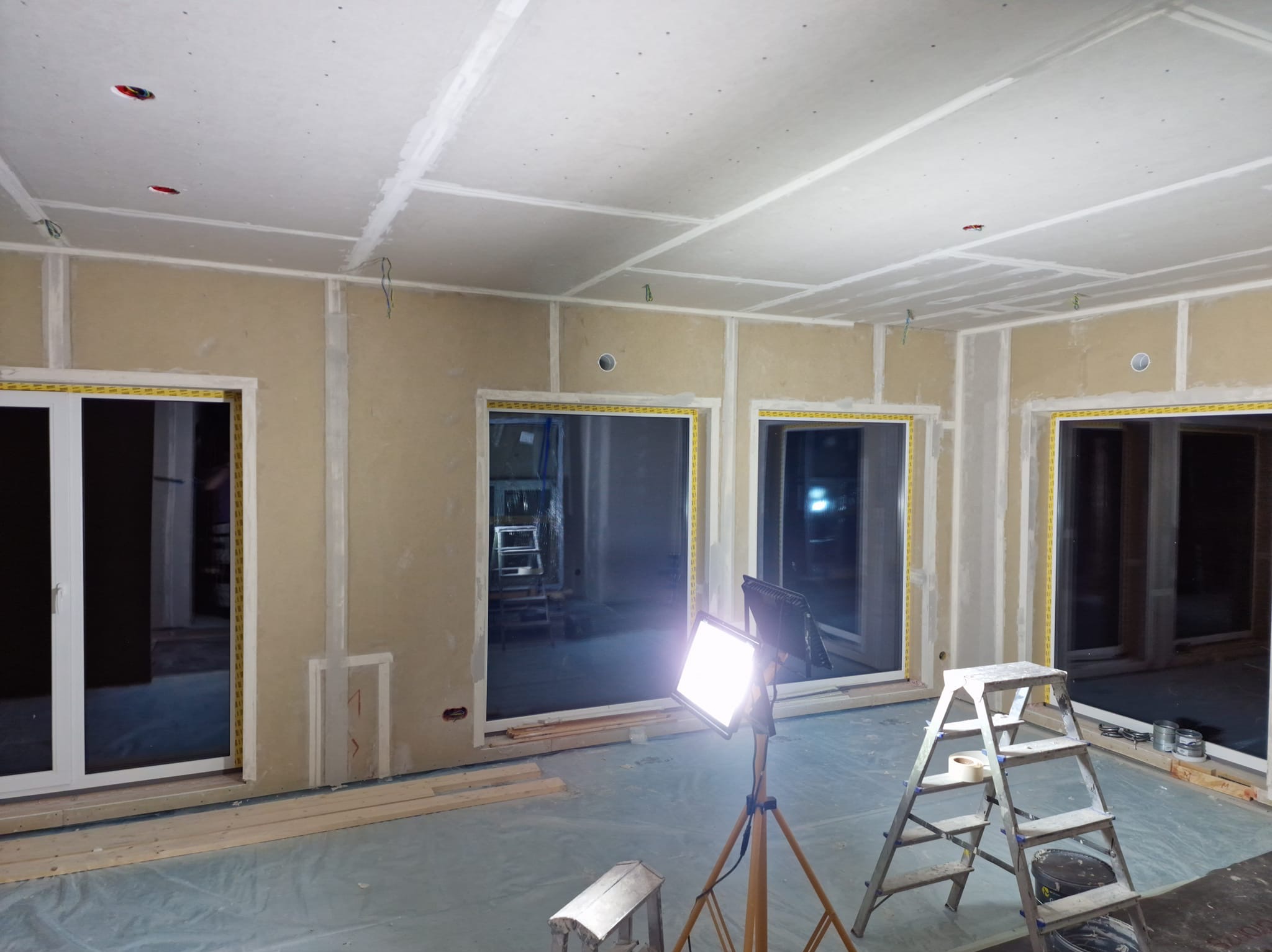 Wnętrze w trakcie prac: ściany z płyt kartonowo-gipsowych, widoczne łączenia, okna z taśmą zabezpieczającą, oświetlenie budowlane i drabina. Podłoga zabezpieczona folią.