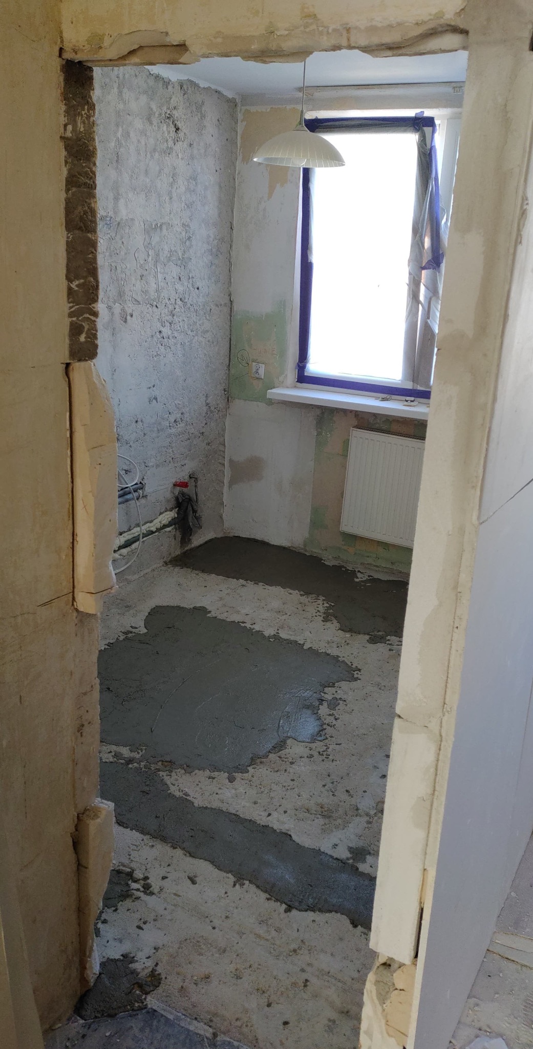 Wnętrze w trakcie prac: częściowo wylana wylewka na podłodze, ściany przygotowane do dalszych prac, widoczne instalacje. Okno z niebieską ramą i grzejnik.
