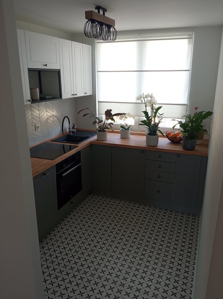 Kompaktowa kuchnia w Kielnie z szarymi szafkami, drewnianym blatem i wzorzystą podłogą. Na blacie rośliny doniczkowe i owoce. Nowoczesne oświetlenie sufitowe.