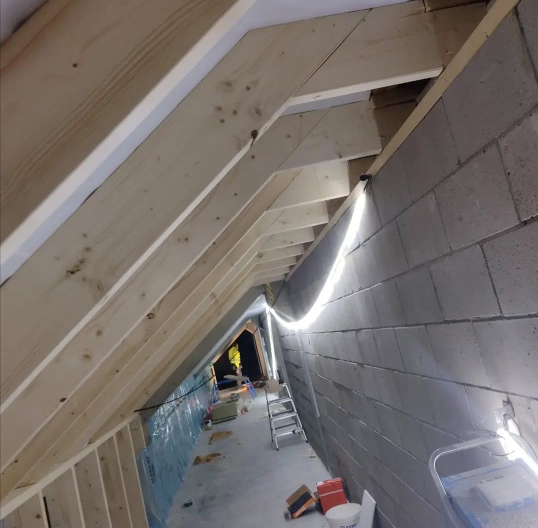 Adaptacja poddasza: drewniana konstrukcja szkieletowa dachu, ściana z bloczków betonowych, oświetlenie LED. Widok w głąb pomieszczenia, drabina i materiały budowlane.