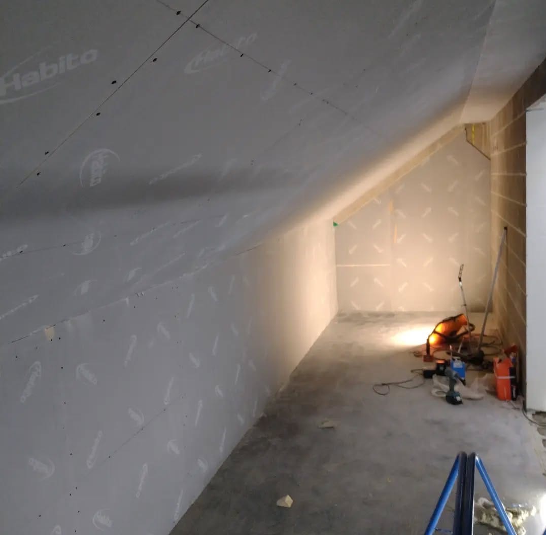 Poddasze w trakcie prac wykończeniowych: ściany i skosy obłożone płytami gipsowymi z widocznym logo producenta, narzędzia pracy w tle, surowa podłoga.