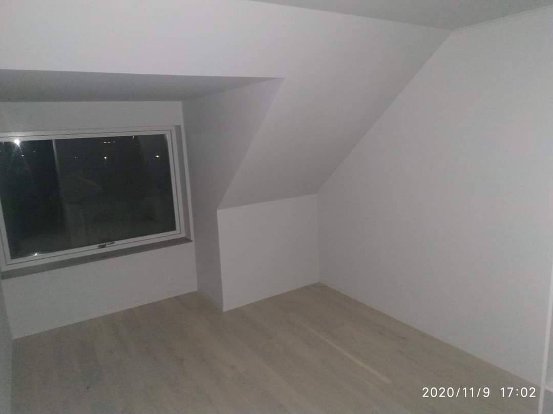 Poddasze po remoncie: białe ściany, jasna podłoga z paneli, okno. Skośny sufit i ściany tworzą unikalny kształt pomieszczenia. Minimalistyczny styl wykończenia.