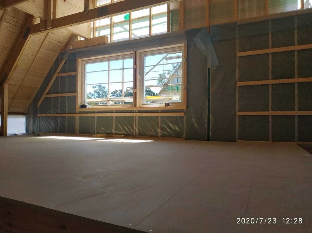 Poddasze w trakcie adaptacji: widoczne ocieplone ściany z drewnianą konstrukcją, okna z widokiem na zewnątrz i surowa podłoga. Prace przygotowawcze do wykończenia.