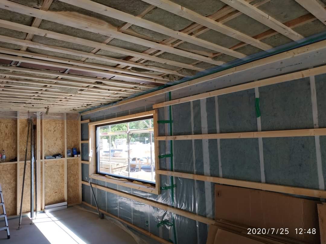 Ocieplenie ścian i sufitu wełną mineralną, widoczne drewniane stelaże i folia paroizolacyjna. Okno w trakcie montażu. Prace wykończeniowe w surowym wnętrzu.