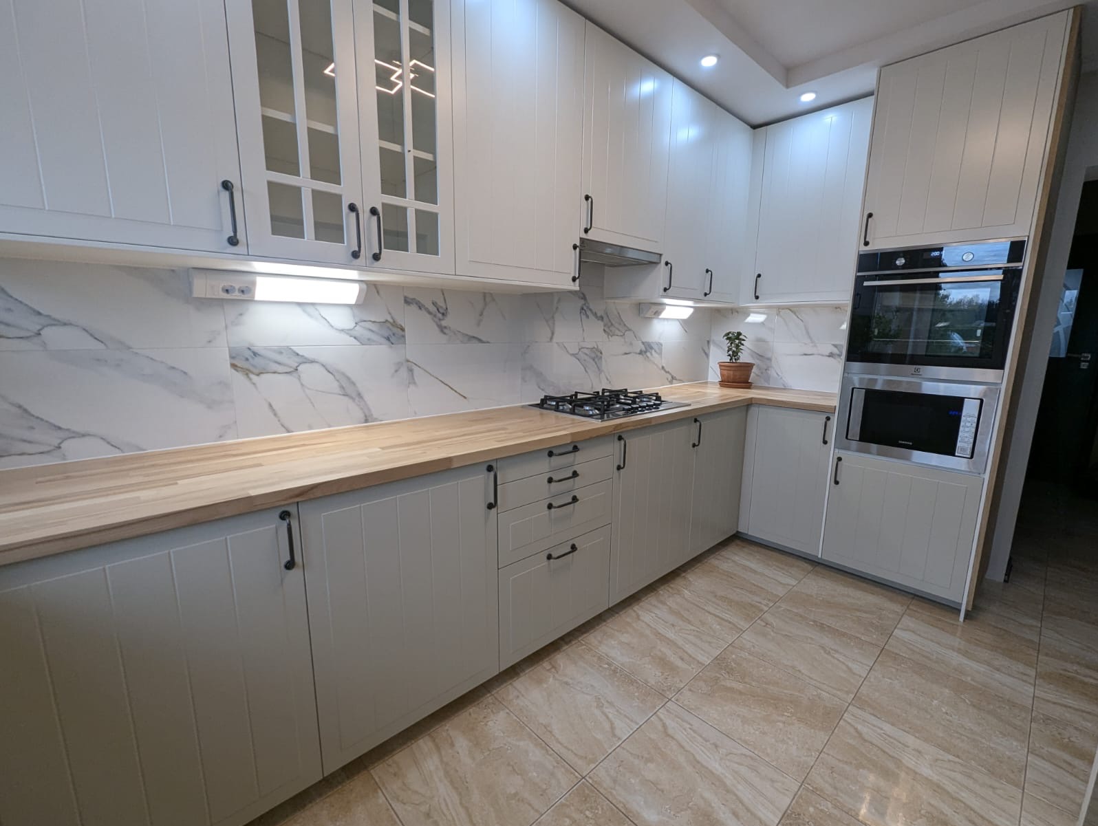 Jasna kuchnia w stylu klasycznym z drewnianym blatem, marmurowymi płytkami i czarnymi uchwytami. Widoczna płyta gazowa i zabudowany piekarnik.