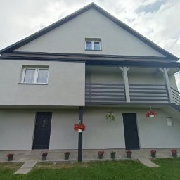 WarmTech Technologies - Dwupiętrowy dom po ociepleniu i malowaniu w odcieniach szarości z czarnymi detalami, kwiaty w doniczkach wzdłuż ścieżki i na balkonie, schody zewnętrzne.