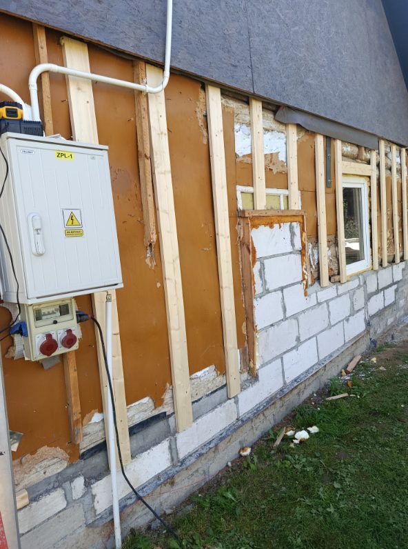 Ściana zewnętrzna domu w trakcie ocieplania: widoczne drewniane listwy, fragmenty starej elewacji, okno i skrzynka elektryczna. Prace przygotowawcze do izolacji.