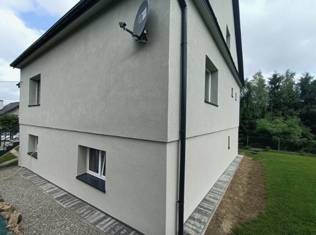 Dwukondygnacyjny dom z szarą elewacją i czarną rynną, widziany z perspektywy ogrodu. Na ścianie antena satelitarna. Nowoczesny styl.