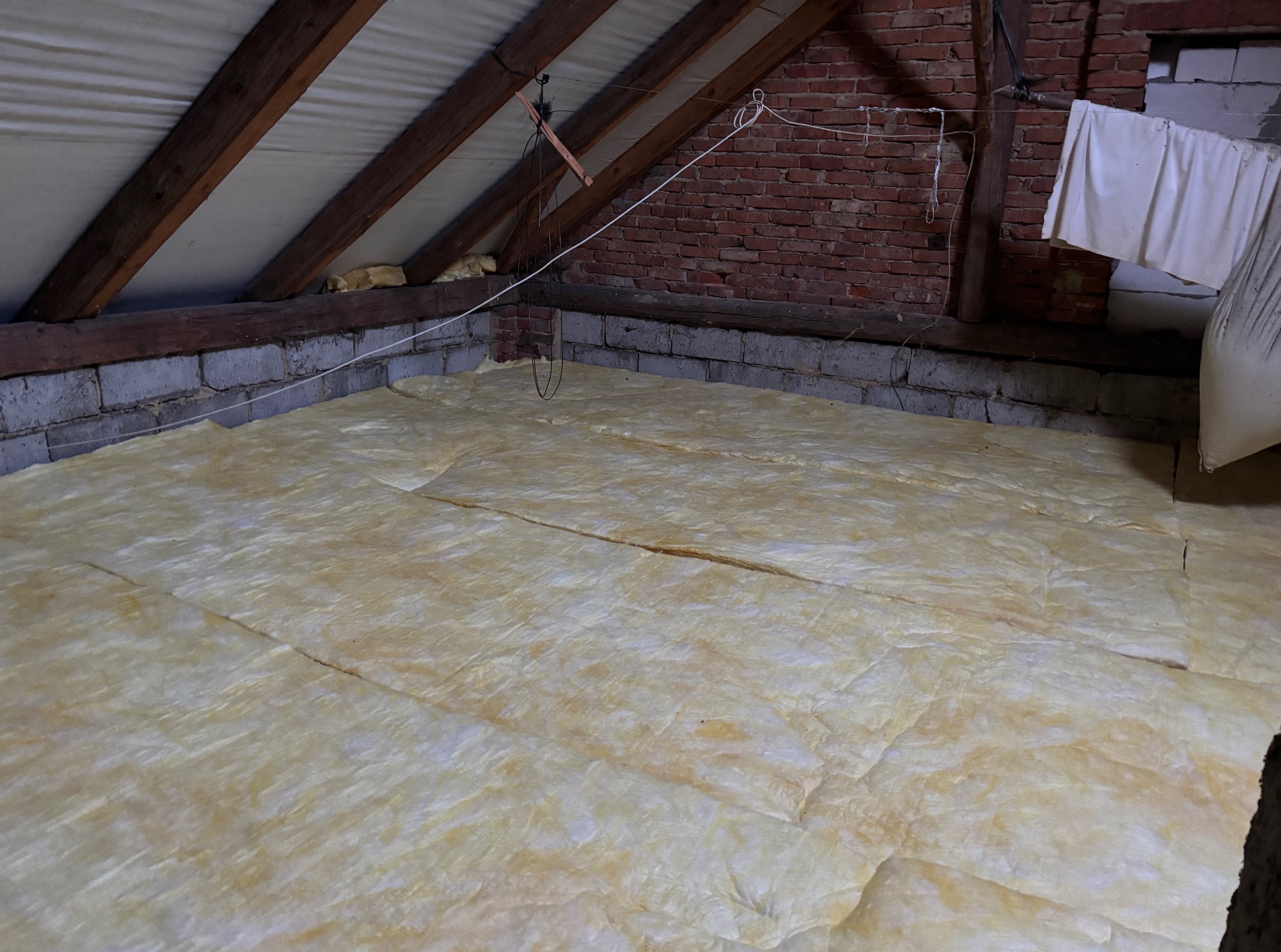 Poddasze z izolacją z wełny mineralnej, widoczne drewniane elementy konstrukcji dachu i fragment muru z cegły. Izolacja ułożona na podłodze.
