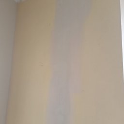 Artur Michalak - Fragment ściany w trakcie szpachlowania. Widoczna jaśniejsza, świeżo nałożona warstwa gładzi na tle ściany w odcieniu kremowym. Górny róg ściany i fragment sufitu.