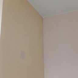Artur Michalak - Pomieszczenie po malowaniu: beżowa ściana z delikatną plamą i jasnoróżowa ściana, biały sufit. Widoczny róg pokoju i fragment okna.