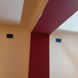 Artur Michalak - Pomarańczowa ściana z czarnym gniazdkiem, bordowa kolumna i biały sufit z listwami. Widoczne taśmy malarskie zabezpieczające gniazdko na ścianie.