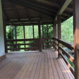 Zadaszony taras drewniany z balustradami, widok na zieleń. Brązowe deski i konstrukcja. Naturalne światło, spokojna atmosfera.