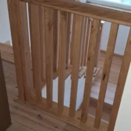 Nowoczesne schody z jasnego drewna z balustradą w minimalistycznym stylu. Widoczne drewniane panele podłogowe i białe ściany. Okno w tle wpuszcza naturalne światło.