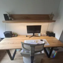 Biurko z blatem z jasnego drewna i metalowymi nogami w kształcie litery V, na tle białej ściany z półką i szafką w skosie, komputer i akcesoria na blacie.