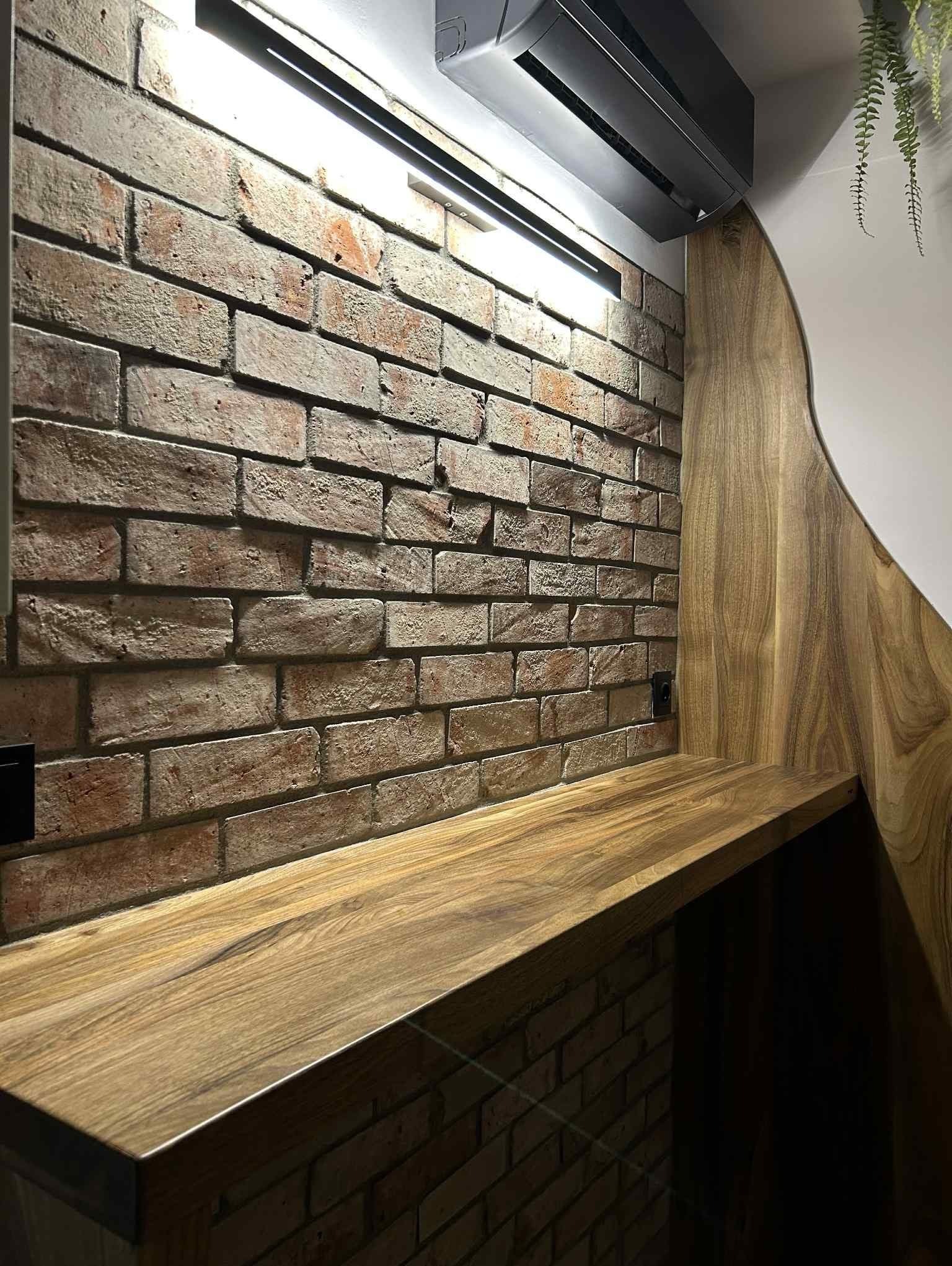 Loftowy detal: ceglana ściana z drewnianym blatem i klimatyzatorem. Widoczne gniazdko elektryczne i fragment drewnianej boazerii. Oświetlenie punktowe.
