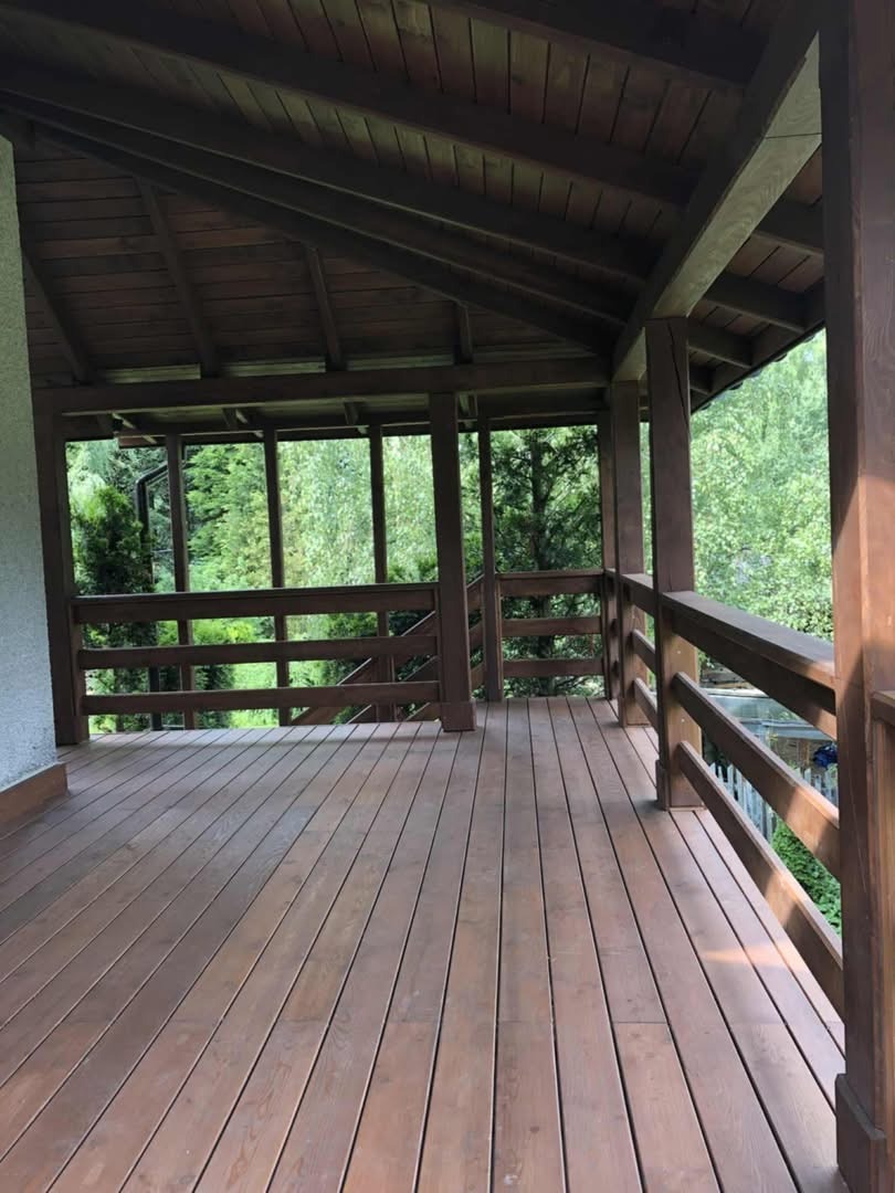 Zadaszony taras drewniany z balustradami, widok na zieleń. Brązowe deski i konstrukcja. Naturalne światło, spokojna atmosfera.