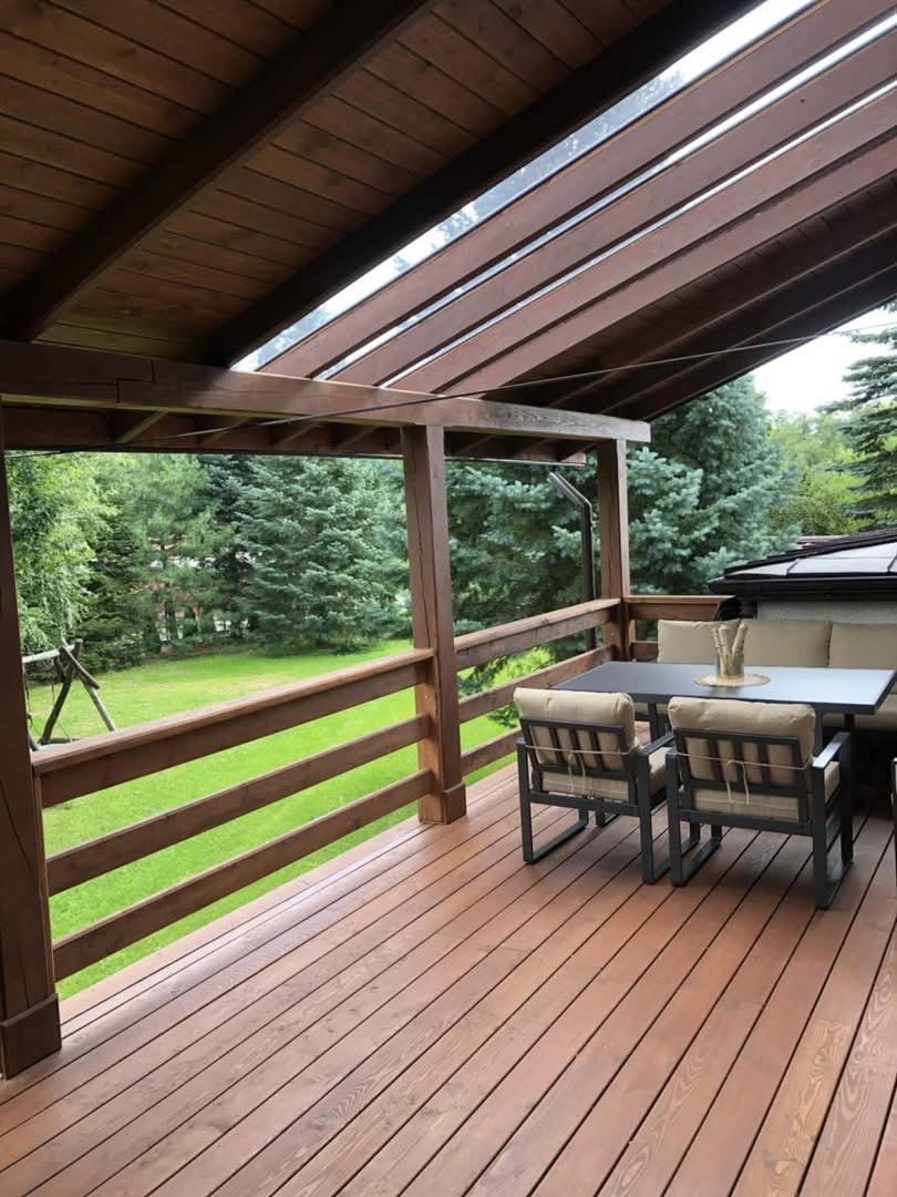 Zadaszony taras z drewnianą podłogą i balustradą. Na tarasie stół z krzesłami i kanapą. Widok na zielony trawnik i drzewa. Drewniana konstrukcja z zadaszeniem.