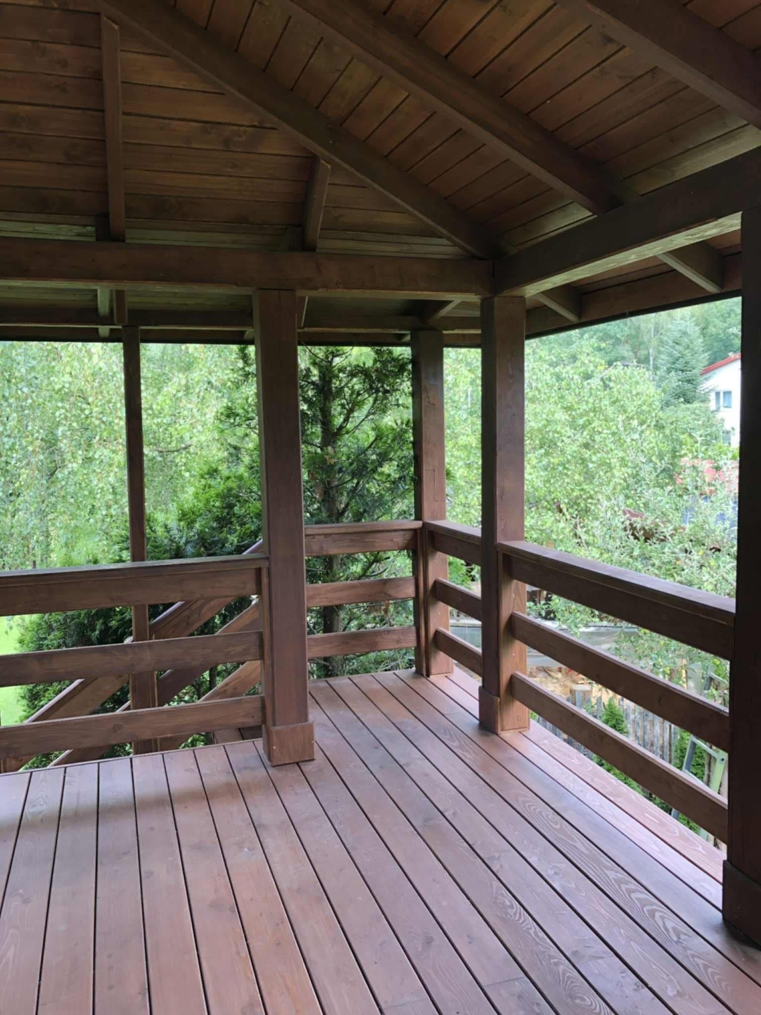 Zadaszony taras z brązowego drewna. Widok na zieleń przez drewniane balustrady. Solidna konstrukcja, naturalne otoczenie, precyzyjne wykonanie.