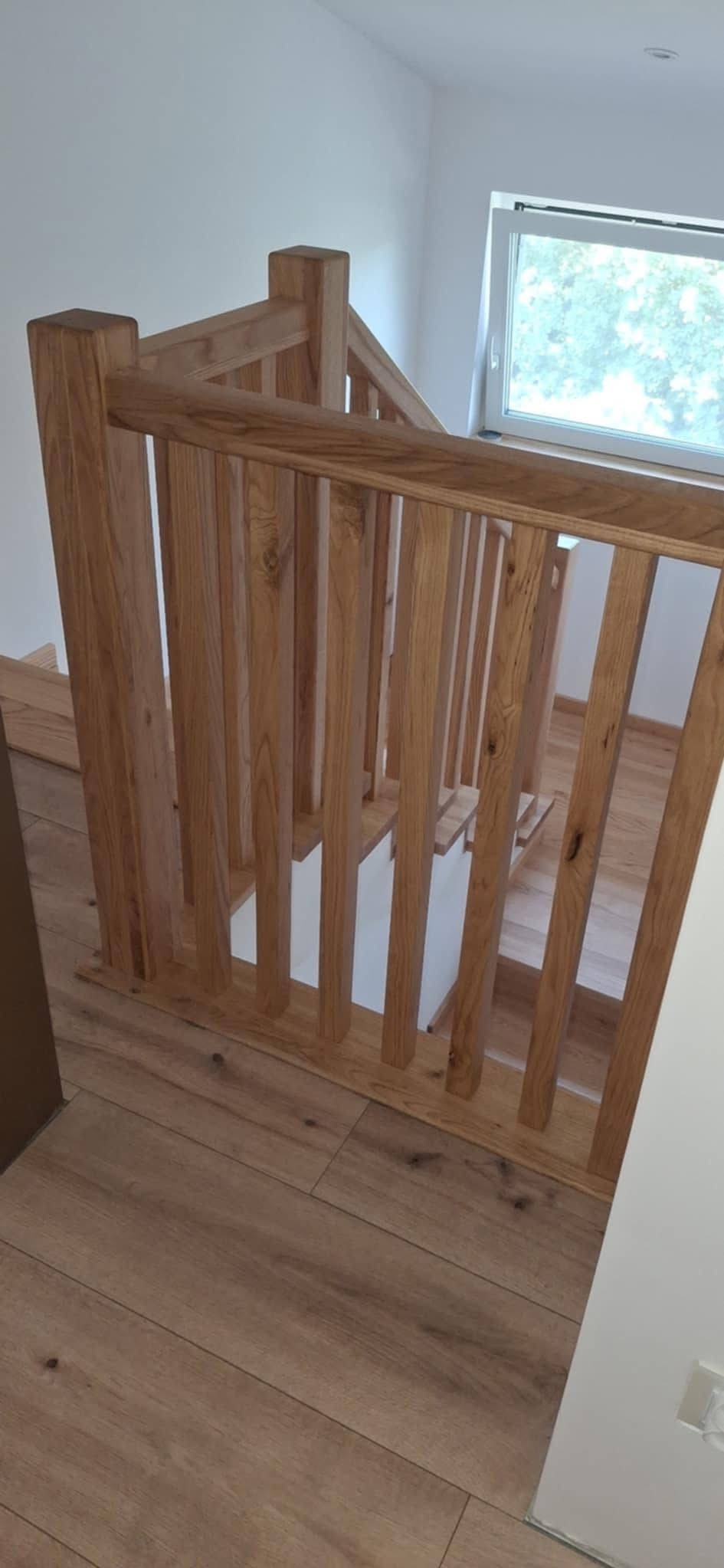 Nowoczesne schody z jasnego drewna z balustradą w minimalistycznym stylu. Widoczne drewniane panele podłogowe i białe ściany. Okno w tle wpuszcza naturalne światło.