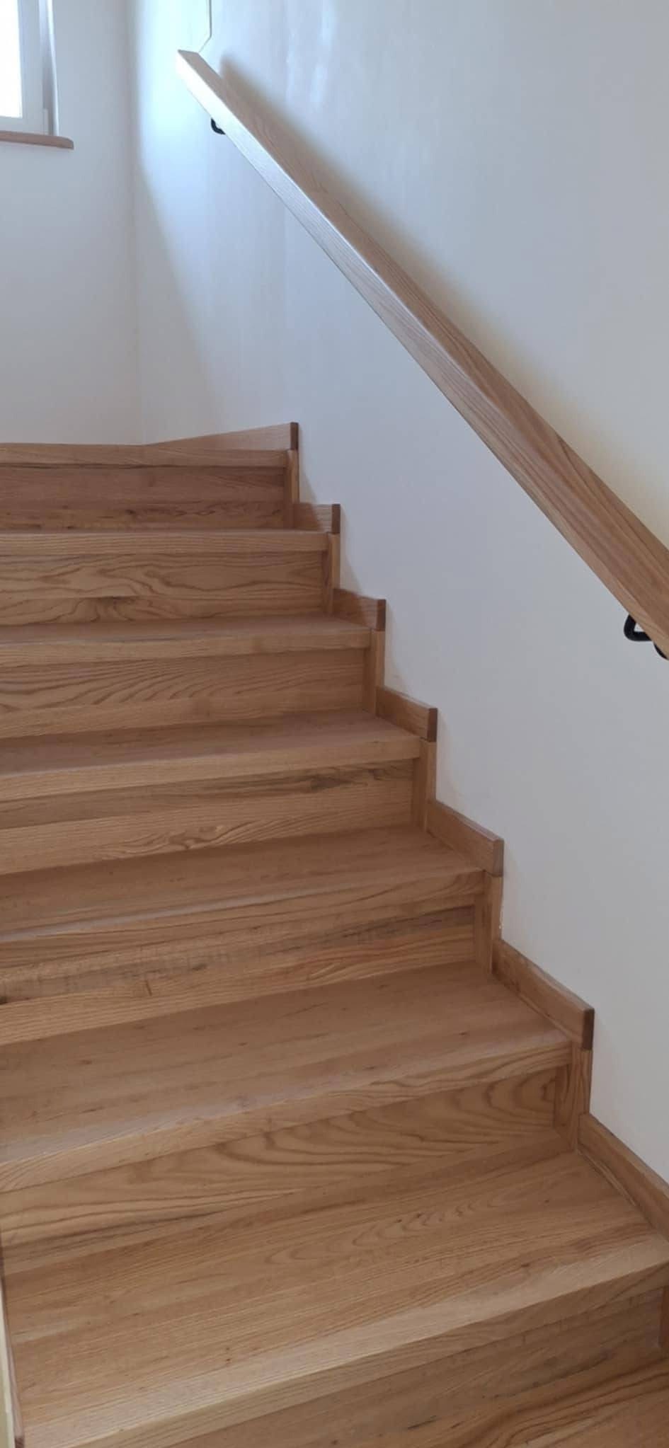 Jasne, drewniane schody z minimalistyczną balustradą przy białej ścianie. Widoczne detale wykończenia stopni i poręczy.