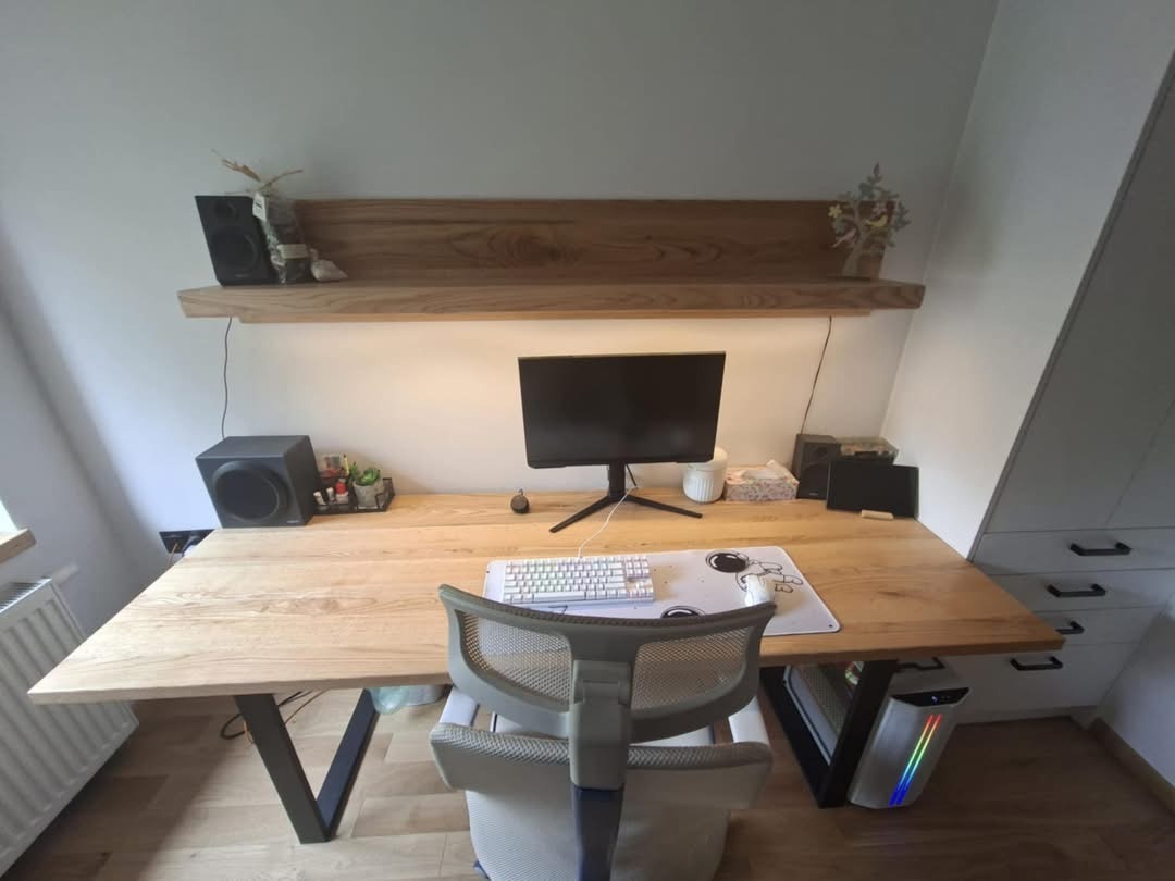 Biurko z blatem z jasnego drewna i metalowymi nogami w kształcie litery V, na tle białej ściany z półką i szafką w skosie, komputer i akcesoria na blacie.