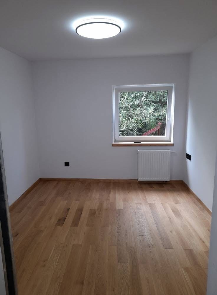 Jasny parkiet w pustym pokoju z oknem na zieleń. Świeżo pomalowane białe ściany i sufit z okrągłą lampą. Grzejnik pod oknem. Nowoczesne wykończenie wnętrza.