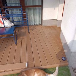 Przemysław Łagodziński P.K-Woods. - Jasnobrązowy taras kompozytowy z widocznymi deskami, niebieskimi krzesłami i psem leżącym na zielonej trawie. Widok na okno z zasłonami i fragment ściany budynku.