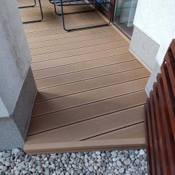 Przemysław Łagodziński P.K-Woods. - Ukończony taras kompozytowy z brązowych desek, z widocznymi niebieskimi krzesłami i kamiennym podłożem. Detal wykończenia przy ścianie budynku.