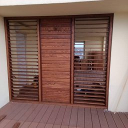 Przemysław Łagodziński P.K-Woods. - Drewniana zabudowa balkonu z lamelami w ciepłym odcieniu brązu, widoczna z zewnątrz na tle jasnej ściany i brązowej podłogi. Widoczne detale konstrukcyjne.