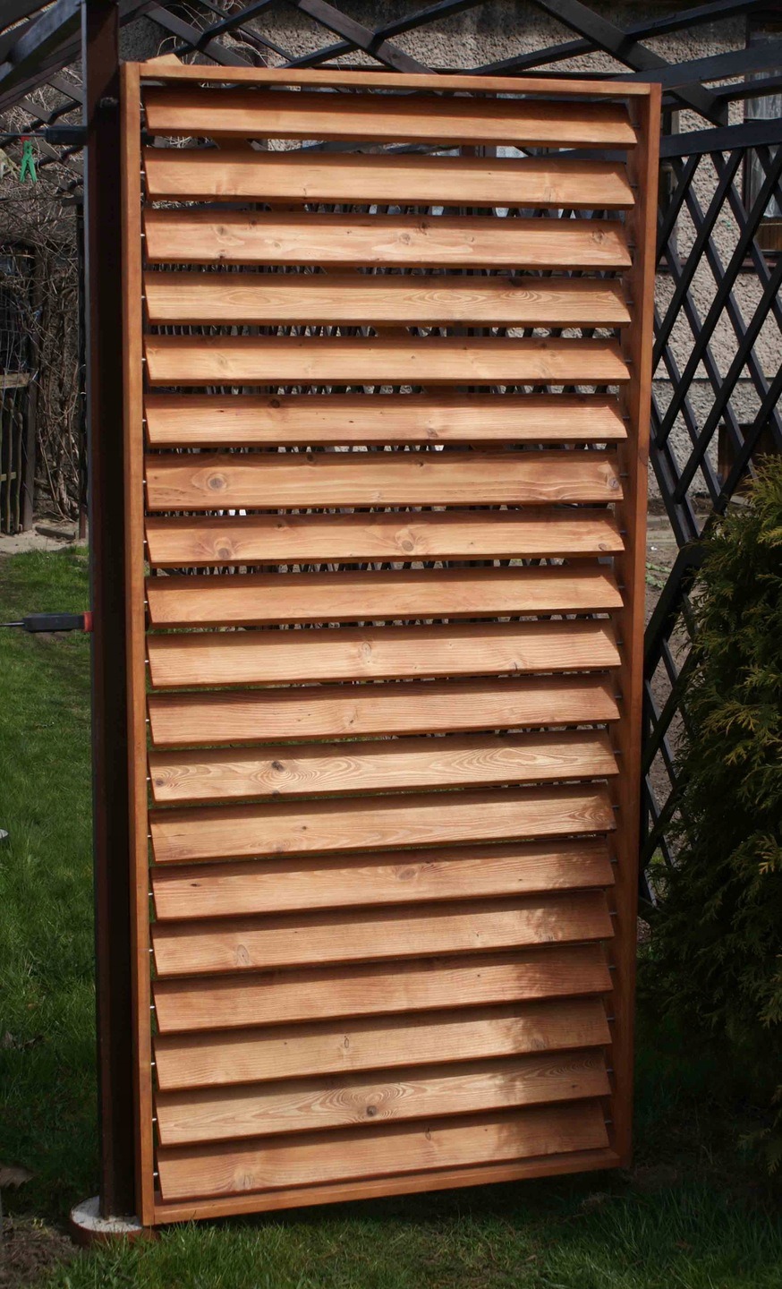 Drewniana, ażurowa ścianka działowa z poziomych listew, ustawiona na trawniku. Widoczna konstrukcja metalowa w tle i fragment roślinności.