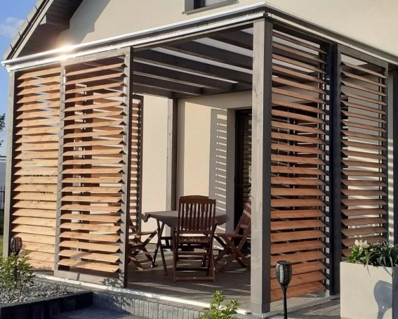 Nowoczesna pergola z drewnianymi żaluzjami, zacieniająca taras z meblami ogrodowymi. Widoczne detale konstrukcyjne i oświetlenie solarne.
