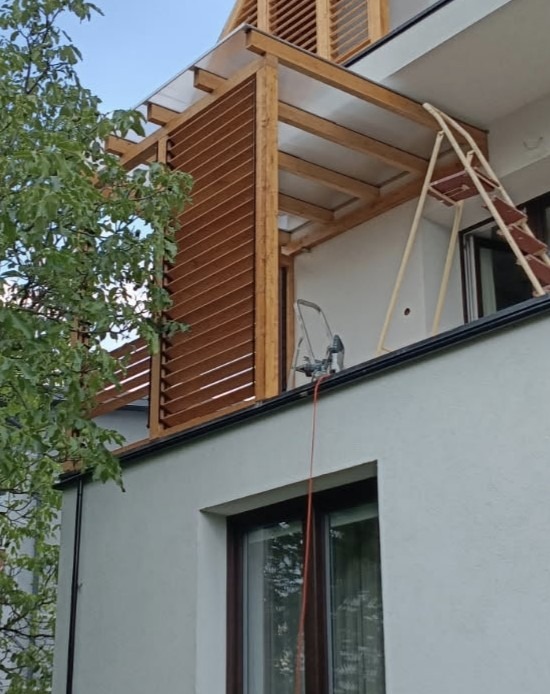Drewniana konstrukcja z lamelami na balkonie, widok z dołu. Na balkonie drabina i narzędzia. Elementy konstrukcji zadaszone przezroczystym materiałem.