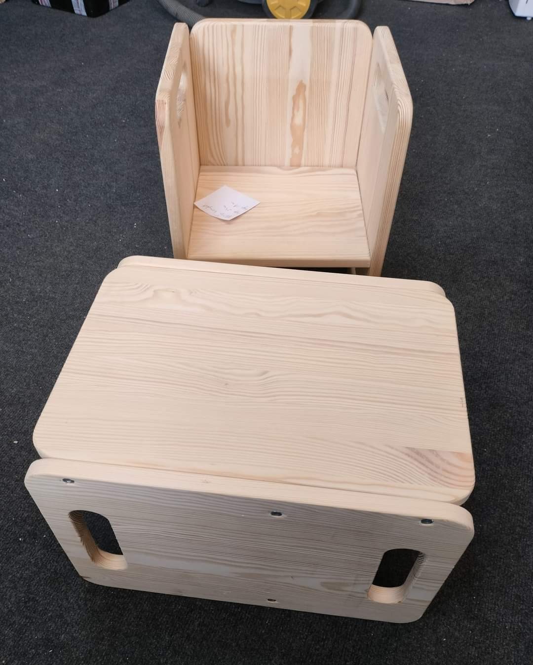 Drewniany, surowy stołek i krzesełko dziecięce z jasnego drewna, z uchwytami. Widoczne ślady po wkrętach. Na siedzisku krzesła leży kartka.