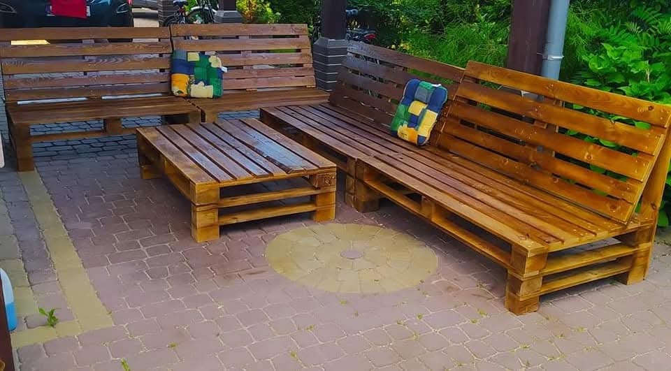 Komplet mebli ogrodowych z palet: dwie ławki z oparciami i stolik kawowy. Całość w ciepłym odcieniu brązu, z kolorowymi poduszkami. Ułożone na kostce brukowej.