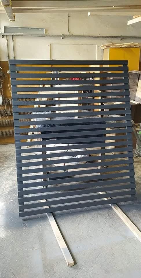 Szara, drewniana konstrukcja z poziomych listew, oparta na dwóch belkach, w warsztacie. Widoczne narzędzia i materiały w tle, sugerujące etap produkcji.