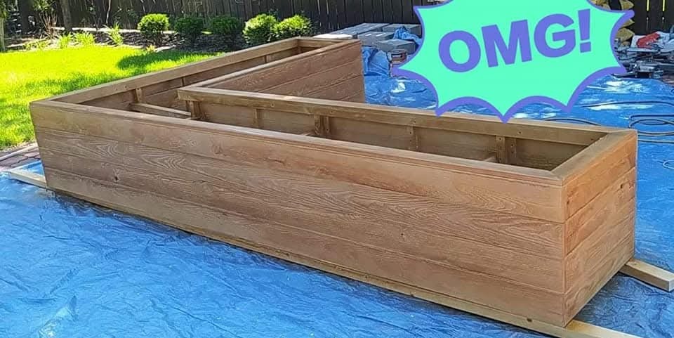 Kanciasta, drewniana donica ogrodowa w kształcie litery L, wykonana z desek o widocznym usłojeniu, ustawiona na niebieskiej folii ochronnej. W tle zielona trawa i krzewy.