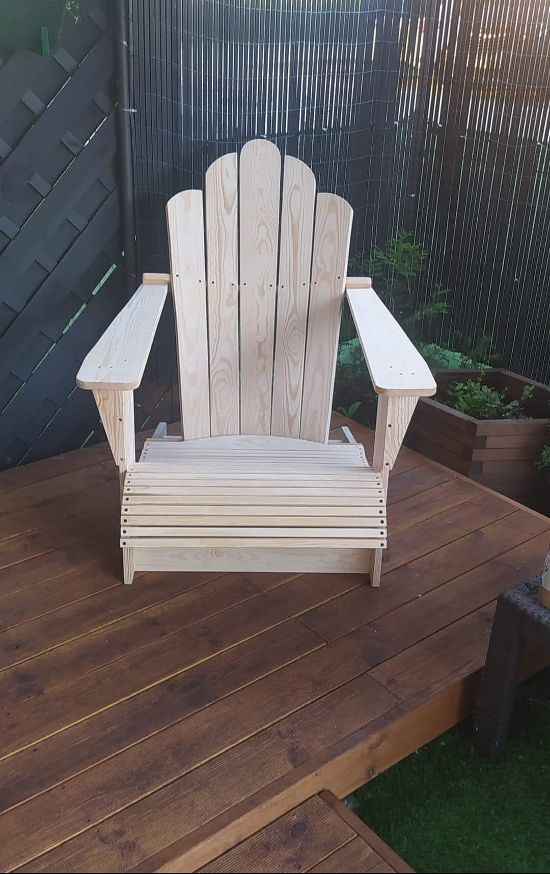 Surowe, drewniane krzesło Adirondack na tarasie z desek. Widoczne detale konstrukcyjne i naturalna faktura drewna. Tło stanowi ciemne ogrodzenie.