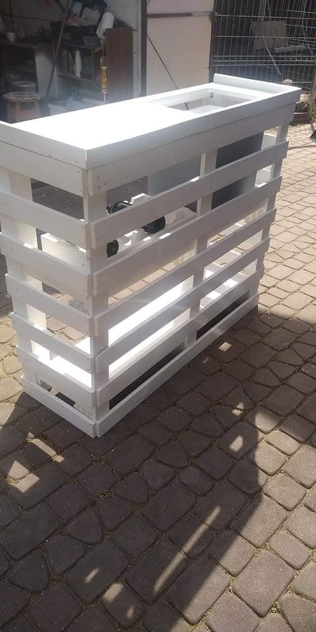 Biała, pomalowana konstrukcja z palet na brukowanym podłożu. Widoczne szczeliny między deskami i cień rzucany przez konstrukcję. W tle zarys budynku i metalowe ogrodzenie.