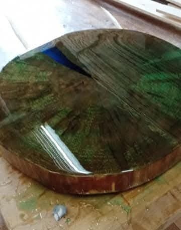 Okrągły blat stołu z drewna i żywicy epoksydowej w odcieniach zieleni i brązu, z widocznymi słojami drewna i fragmentem niebieskiej żywicy. Wykończenie na wysoki połysk.