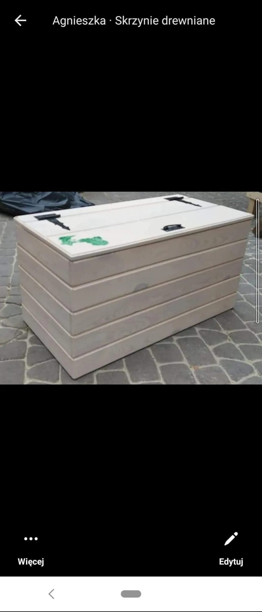 Drewniana skrzynia ogrodowa z poziomych desek, z czarnymi zawiasami, na kostce brukowej. Widoczny fragment czarnej folii w tle. Jasne, naturalne światło.