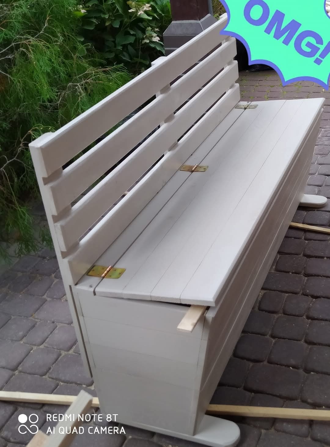 Biała, drewniana ławka z oparciem i schowkiem, widoczna na tle zieleni i kostki brukowej. Zawiasy w kolorze mosiądzu. Etap montażu, widoczne listwy pod ławką.