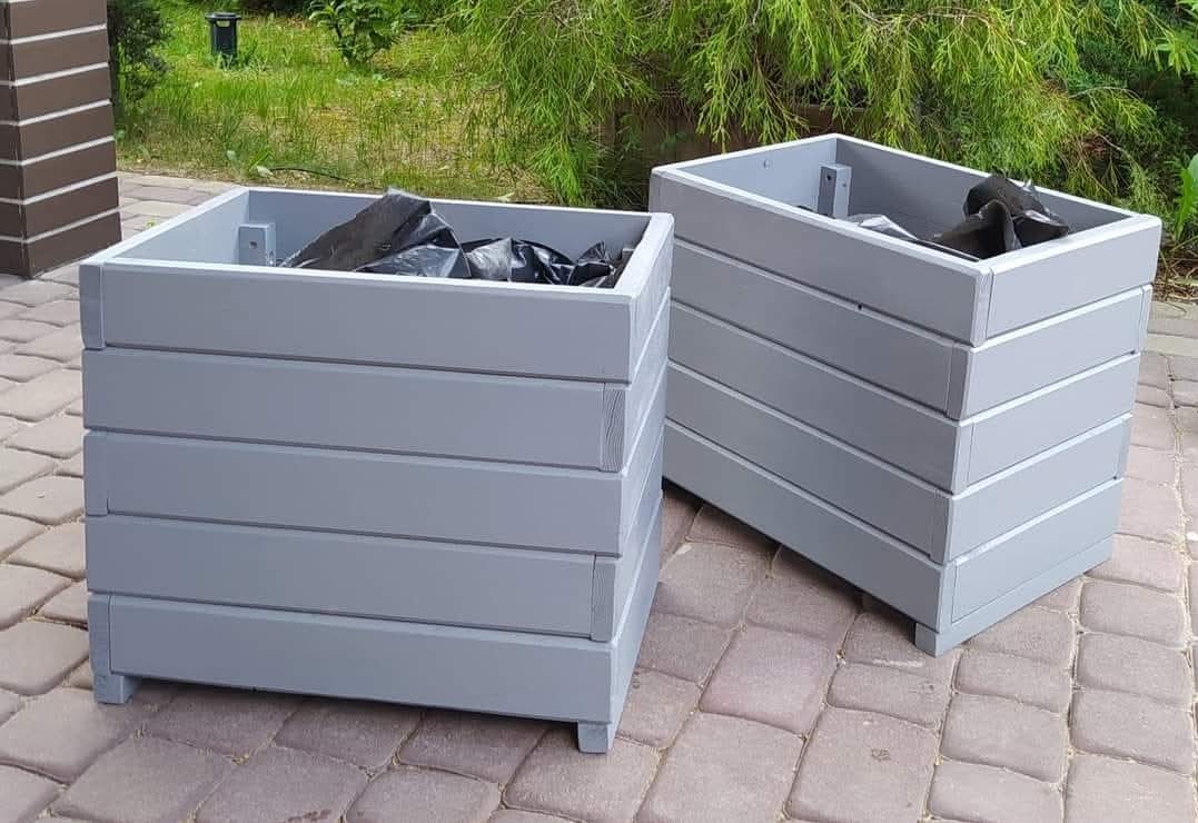 Dwie szare, drewniane donice na tarasie z kostki brukowej. Donice w stylu minimalistycznym, wypełnione czarną folią. Widoczne elementy konstrukcyjne.