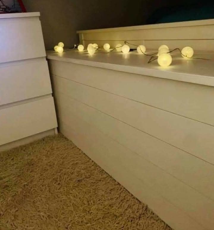 Biała zabudowa z poziomych desek, oświetlona girlandą kulkowych lampek, obok komoda i dywan z długim włosiem. Ciepłe, przytulne wnętrze.