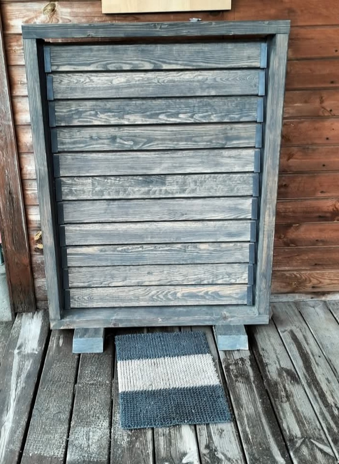 Drewniana, szara przesłona z poziomych listew na tle drewnianej ściany. U dołu leży wycieraczka w paski. Widoczne detale konstrukcyjne i faktura drewna.
