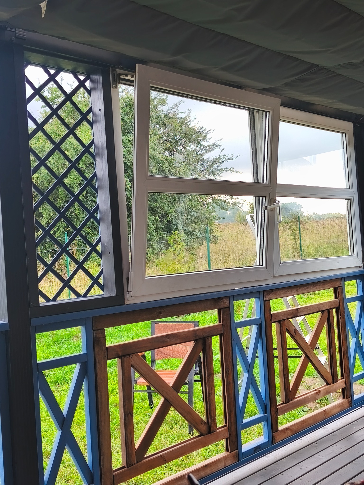 Zabudowa werandy z drewnianą balustradą w kolorach brązu i błękitu, okno PCV z widokiem na zieleń. Kratka dekoracyjna z tyłu. Widok z zewnątrz.