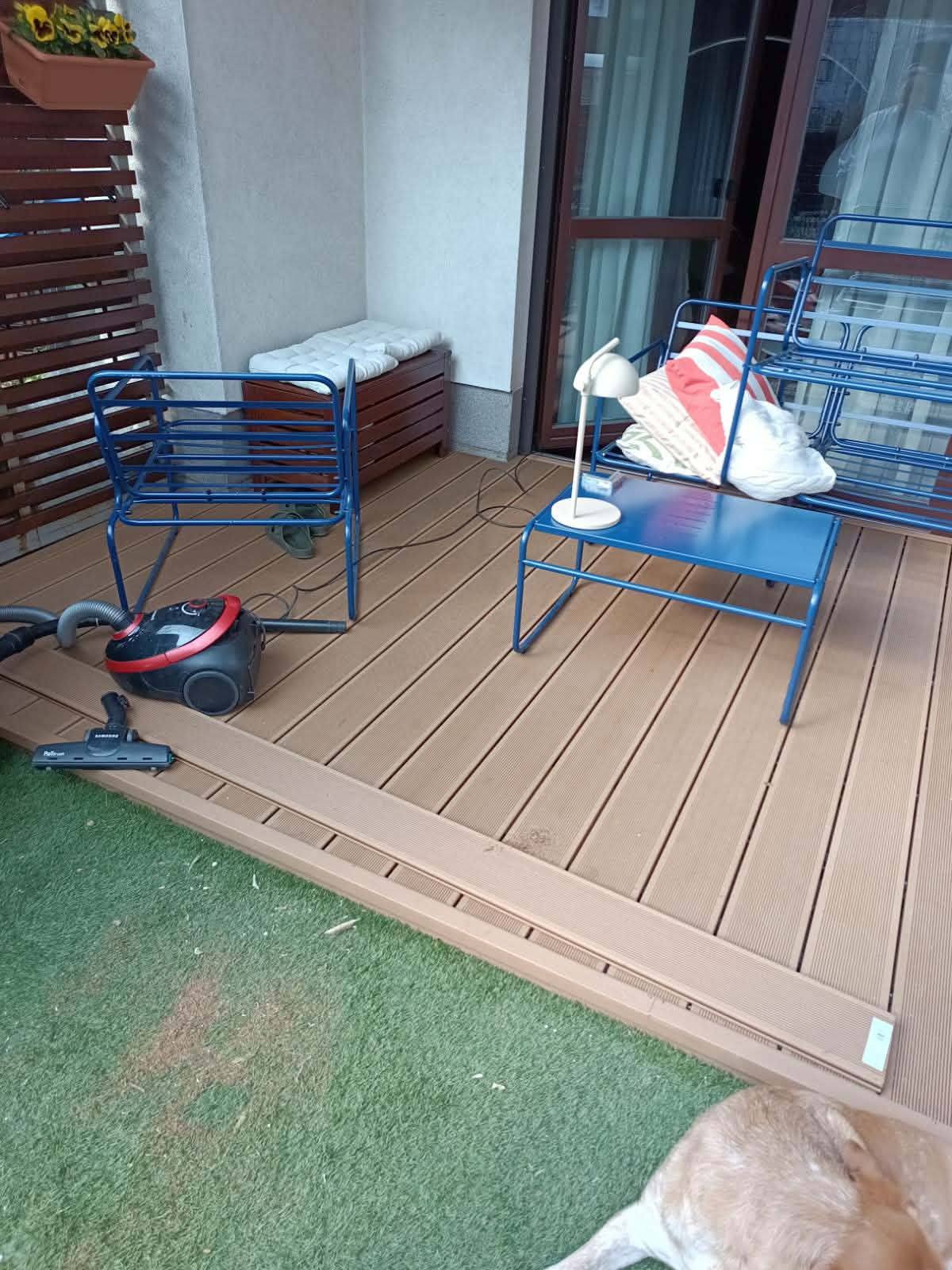 Jasny taras kompozytowy z niebieskimi meblami ogrodowymi i odkurzaczem. Widoczne deski tarasowe i sztuczna trawa. Częściowo widoczny pies w prawym dolnym rogu.