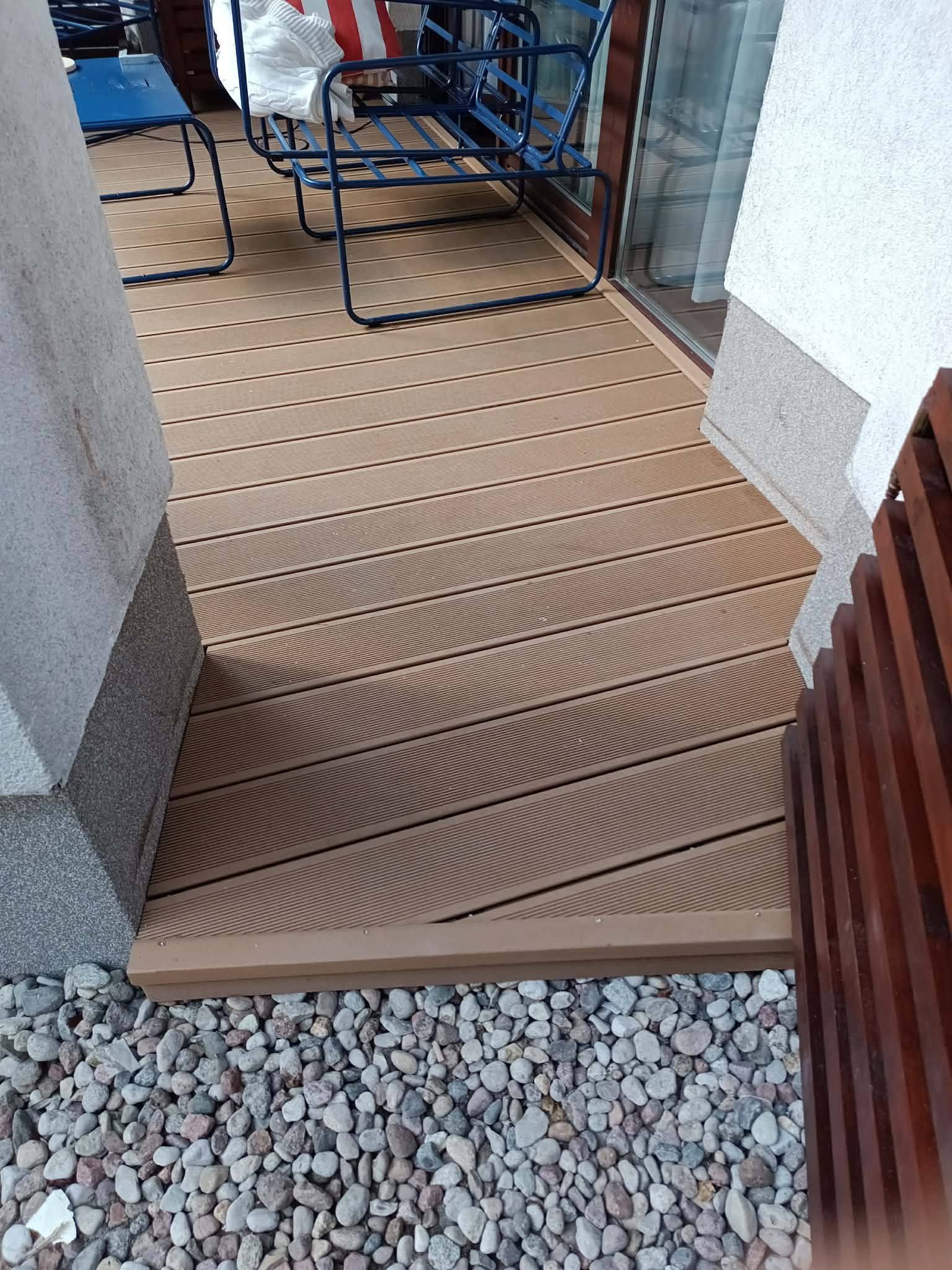 Ukończony taras kompozytowy z brązowych desek, z widocznymi niebieskimi krzesłami i kamiennym podłożem. Detal wykończenia przy ścianie budynku.