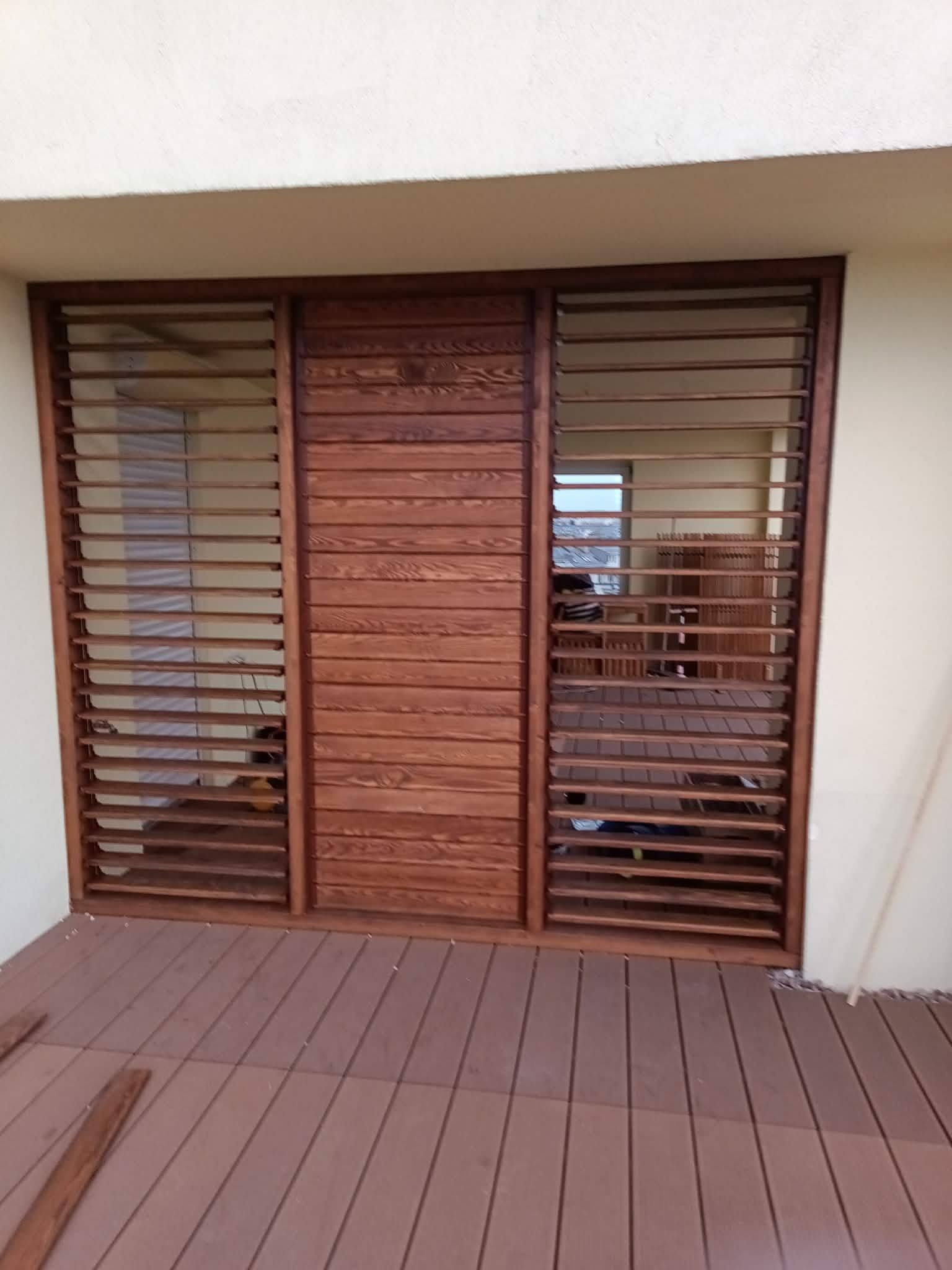 Drewniana zabudowa balkonu z lamelami w ciepłym odcieniu brązu, widoczna z zewnątrz na tle jasnej ściany i brązowej podłogi. Widoczne detale konstrukcyjne.